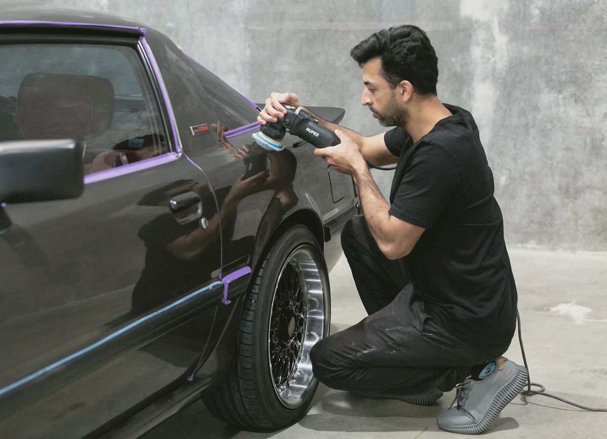 A detailer machine-polishing the door of a dark car, with the trim taped off